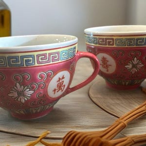 May include: Two red and white ceramic teacups with handles, featuring floral and geometric patterns. The cups have Chinese characters and are set on round wooden coasters. A bottle of oil is in the background.