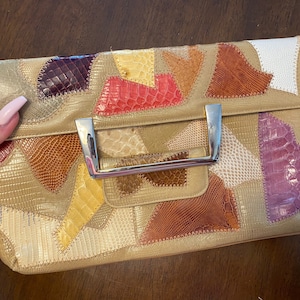May include: A beige clutch with a patchwork design. The clutch features a variety of textured leather pieces in shades of gold, brown, red, and purple. It has a silver metal handle and clasp.