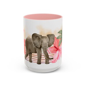 May include: White ceramic mug with a pink interior, featuring a watercolor illustration of a gray elephant. The design includes red flowers and a striped background, creating a tropical theme. The mug is a standard size for hot beverages.