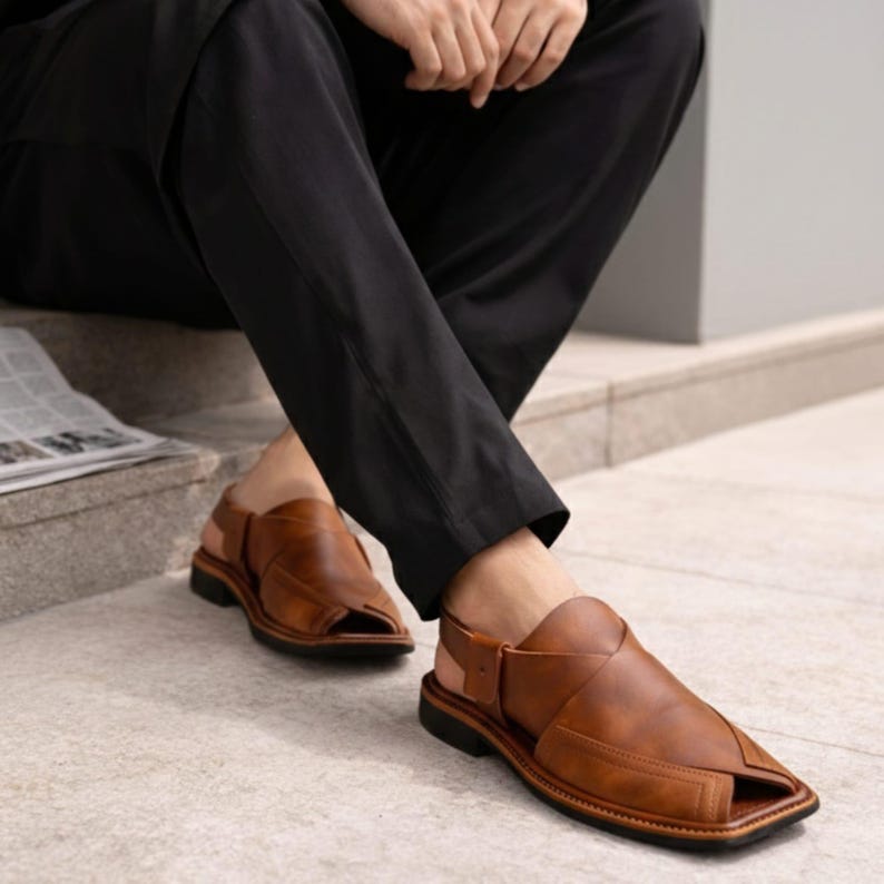 May include: Brown leather sandals with a square toe and a strap around the ankle. The sandals have a dark sole and are worn with black pants. The sandals appear to be new and unworn.