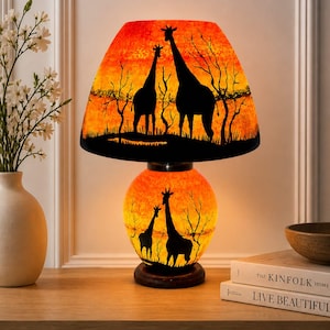 Op de afbeelding: Decoratieve tafellamp met een oranje en gele lampenkap met silhouetten van giraffen tegen een zonsondergang. De voet van de lamp toont ook een giraffensilhouet. De lamp staat op een houten voet.