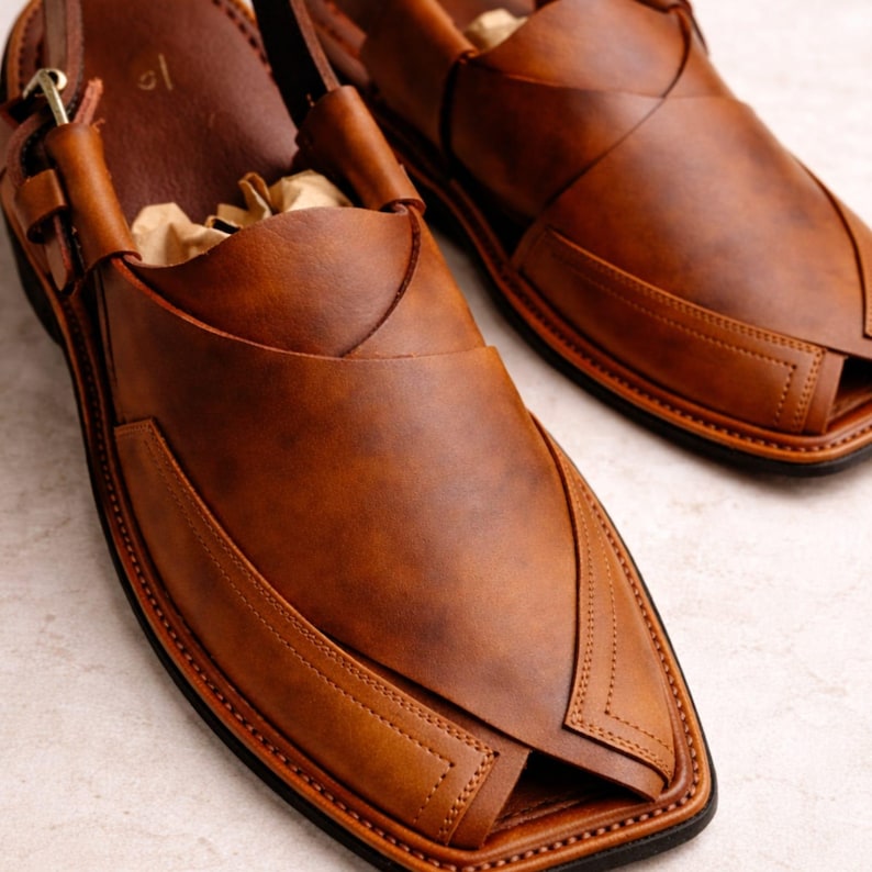 May include: A pair of brown leather sandals with a unique design. The sandals feature a toe opening, a strap that goes around the ankle, and decorative stitching. The leather has a rich, warm tone, and the soles are black.