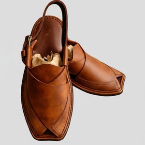 May include: A pair of brown leather Peshawari sandals. These open-toe sandals feature a unique design with a strap that goes around the ankle and a leather upper with decorative stitching. The sandals are filled with brown paper.