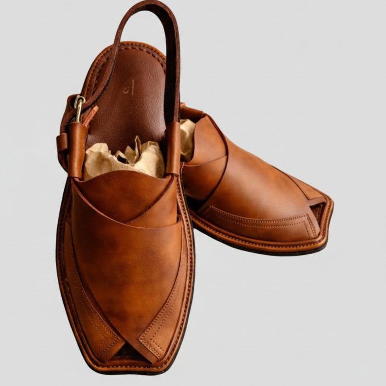 May include: A pair of brown leather Peshawari sandals. These open-toe sandals feature a unique design with a strap that goes around the ankle and a leather upper with decorative stitching. The sandals are filled with brown paper.