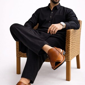 May include: A person wearing a black outfit, including a long-sleeved shirt and pants, is seated in a wicker chair. The person is wearing brown leather sandals. The outfit is suitable for formal or casual wear.