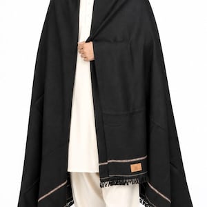 May include: A solid black shawl featuring a thin, multi-colored horizontal stripe detail near the bottom edge, finished with short black fringe. A small rectangular tan brand label is stitched onto the lower corner of the fabric.