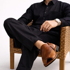 May include: A person wearing a black shirt and pants, seated in a wicker chair, with brown leather sandals. The sandals have a strap around the ankle and a dark sole. The person is also wearing a watch and sunglasses.