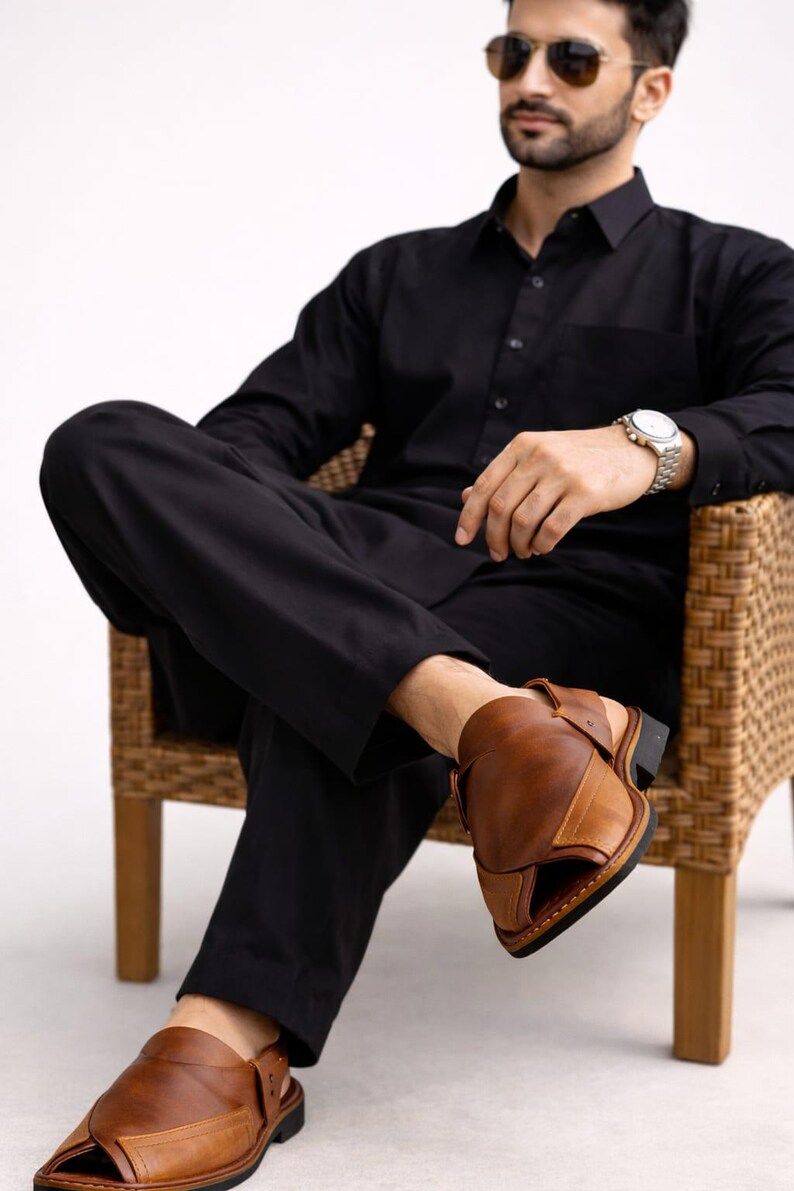 May include: A person wearing a black shirt and pants, seated in a wicker chair, with brown leather sandals. The sandals have a strap around the ankle and a dark sole. The person is also wearing a watch and sunglasses.