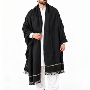 May include: A solid black shawl featuring a thin horizontal gold-toned stripe near the bottom hem and black fringe detailing. The shawl is draped over a white long-sleeved shirt and white trousers, with brown leather shoes visible at the base.