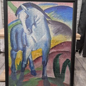 Op de afbeelding: Een ingelijste puzzel met een blauw paard tegen een kleurrijke achtergrond. Het kunstwerk heeft een moderne, abstracte stijl, met het paard als centraal figuur. De puzzel is ingelijst in een zwart frame, klaar om te worden tentoongesteld. Een levendig kunstwerk.