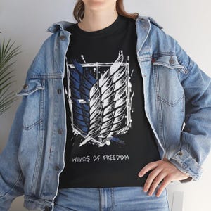 May include: Black t-shirt with a white and blue emblem of stylized wings in a rectangular frame. The text "WINGS OF FREEDOM" is printed below the emblem in white. The shirt is worn with a blue denim jacket and jeans.