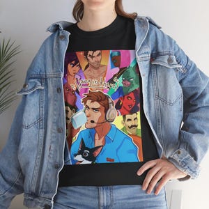 Könnte beinhalten: Schwarzes T-Shirt mit einem farbenfrohen Grafikdesign mit dem Text "Hang in there!". Das Design zeigt Cartoon-Charaktere, einen Mann, der aus einer Tasse trinkt, und einen kleinen Hund. Das Shirt wird mit einer Jeansjacke getragen.