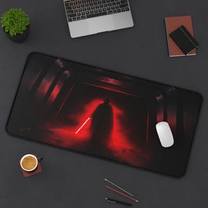 May include: Black rectangular mouse mat with a red and black graphic of a cloaked figure with a red lightsaber in a dark, smoky setting. A white computer mouse is on the right. A laptop, pencils, and a coffee cup are also visible.