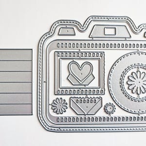 May include: A silver metal die cut in the shape of a camera with a rectangular die cut to the left. The camera die cut features a heart, flower, and envelope design. The camera has a lens, viewfinder, and decorative details.