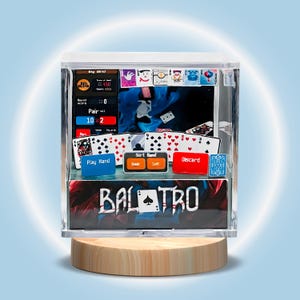 May include: A clear acrylic display case with a poker theme. The case shows playing cards, game interface elements, and the word "BALOTRO." It rests on a round wooden base, set against a blue and white backdrop.