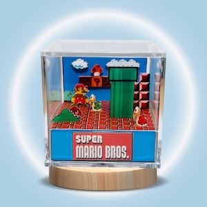 May include: A Super Mario Bros. diorama in a clear acrylic box. The scene includes Mario, a green pipe, and other game elements. The base is light-coloured wood. The text "SUPER MARIO BROS." is on a red sign.