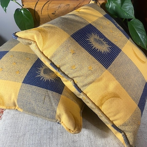 Könnte beinhalten: Zwei dekorative Kissen mit einem gelb-marineblauen Karomuster. Die Kissen zeigen Himmelsmotive von Sonne, Mond und Sternen. Die Kissen sind mit einer marineblauen Paspel versehen.