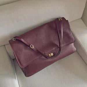 May include: A deep burgundy shoulder bag with a textured surface, featuring a large front flap, thin adjustable shoulder straps, and a gold-toned rectangular turn-lock closure. The bag has a structured rectangular shape with rounded corners.
