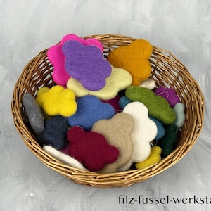 Può includere: Un cesto di vimini pieno di forme di nuvole di feltro colorate, tra cui rosa, viola, giallo, blu e bianco. Il cesto è appoggiato su una superficie grigio chiaro. L'indirizzo del sito web "filz-fussel-werkstatt.de" è visibile.