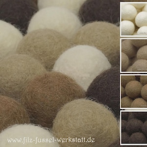 Könnte beinhalten: Nahaufnahme einer Ansammlung von Filzkugeln in verschiedenen neutralen Farben, darunter Creme, Beige, Braun und Dunkelgrau. Die Kugeln sind dicht gepackt und bilden eine strukturierte Oberfläche. Die Website-Adresse www.filz-fussel-werkstatt.de ist sichtbar.