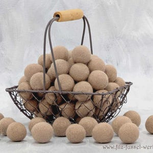 Könnte beinhalten: Ein Drahtkorb gefüllt mit zahlreichen beigefarbenen Filzkugeln, einige davon liegen am Boden. Der Korb hat einen Holzgriff und einen dunklen Metallrahmen. Die Filzkugeln haben eine einheitliche Größe und Farbe und erzeugen eine natürliche Textur.
