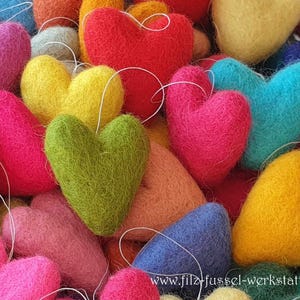 Könnte beinhalten: Eine Sammlung bunter Filzherz-Ornamente in Rot-, Rosa-, Gelb-, Grün- und Blautönen. Jedes Herz hat eine weiße Schnur zum Aufhängen. Das Bild zeigt eine Vielzahl von herzförmigen Dekorationen.