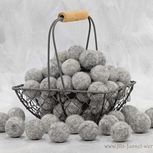 Könnte beinhalten: Ein Drahtkorb gefüllt mit grauen Filzkugeln, einige davon liegen am Boden. Der Korb hat einen Holzgriff. Die Filzkugeln haben eine neutrale graue Farbe, und die Gesamtästhetik ist rustikal und natürlich.