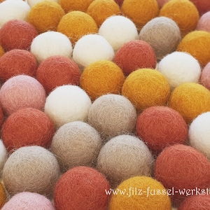 Könnte beinhalten: Nahaufnahme einer Sammlung farbenfroher Filzkugeln in Weiß-, Rosa-, Orange-, Gelb- und Grautönen. Die Kugeln sind eng aneinander angeordnet und bilden eine strukturierte Oberfläche. Die Filzkugeln werden wahrscheinlich für Bastelarbeiten oder Heimdekoration verwendet.