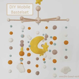 Op de afbeelding: Een doe-het-zelf mobiel kit met een houten frame, met vilten ballen in wit, oranje en bruin, samen met een gele maan en ster. De tekst "DIY Mobile Bastelset" is zichtbaar.