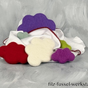 Może przedstawiać: Kolekcja filcowych ozdób w kształcie chmur w różnych kolorach, w tym czerwonym, fioletowym, białym i zielonym, ułożonych na białej tkaninie z czerwoną lamówką. Tekst "filz-fussel-werkstatt.de" jest widoczny na dole.