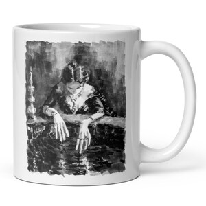 May include: White ceramic mug featuring a black and white illustration of a woman in a bathtub. The artwork depicts a woman with long hair, jewelry, and a candle. The mug has a white handle and a glossy finish.