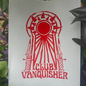 May include: A white rectangular print featuring a red ink block-style illustration of a keyhole shape containing a sunburst motif, flanked by two vertical daggers. Below the graphic, the text CLUB VANQUISHER is printed in a bold red font.