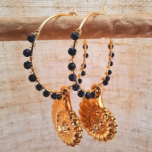 Puede incluir: Pendientes de aro dorados con cuentas negras y un colgante en forma de concha. Los pendientes están suspendidos de una clavija de madera sobre un fondo neutro. Los pendientes tienen un diseño en espiral.