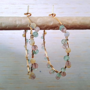 May include: A pair of gold-colored hoop earrings. Each earring is adorned with a series of small, multi-colored beads in shades of pink, green, and yellow. The earrings are suspended from a wooden dowel.