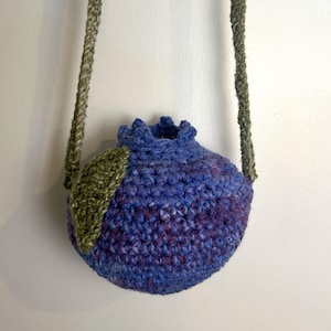 Op de afbeelding: Een handgemaakte gehaakte crossbodytas in de vorm van een bosbes. De tas is voornamelijk blauw met paarse accenten, een groen blad en een groene band. De tas hangt tegen een witte achtergrond.