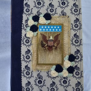 May include: A rectangular journal with a cream-colored fabric cover featuring a repeating navy blue pattern of flags and stars. The cover is decorated with a central rectangular patch showing a bald eagle, the text 1776, and fabric rosettes.