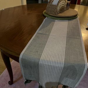 May include: A gray and white table runner with a striped design. The runner is made of a woven fabric and has a central white stripe with dotted accents. The runner is placed on a wooden table.