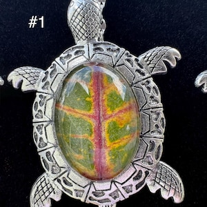 May include: A silver-toned turtle pendant necklace. The pendant features a large, oval-shaped stone with green, yellow, and red patterns. The turtle's shell and head are detailed with textured designs. The necklace is displayed against a black background.