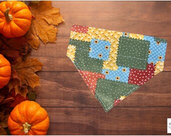 Op de afbeelding: Een patchwork honden bandana met een herfstthema, met vierkanten van groene, blauwe, gele en rode stoffen. De bandana ligt op een houten oppervlak met pompoenen, herfstbladeren en kaneelstokjes.