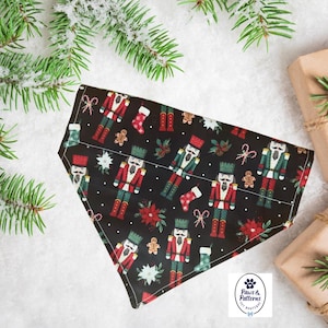 May include: A black dog bandana with a Christmas-themed pattern featuring nutcrackers, stockings, and poinsettias. The bandana is triangular and lies on a snowy surface, next to wrapped gifts and evergreen branches.