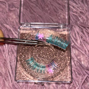May include: A clear, hinged case containing a set of false eyelashes. The lashes are teal and pink with glitter accents. A pair of tweezers holds one lash. The case is filled with glitter and rests on a pink, fuzzy surface.