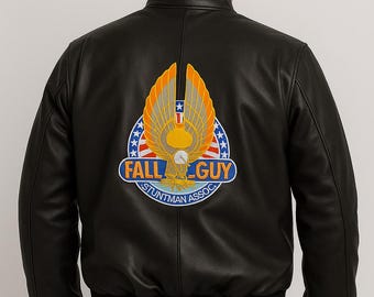 May include: Black leather bomber jacket with a ribbed collar and cuffs. The back features a colorful patch with an eagle, wings, and the text "FALL GUY STUNTMAN ASSOC" in a circular design. The jacket is worn over dark blue jeans.