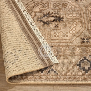 May include: A tan area rug with a folded corner revealing a light beige underside and white fringe. The rug features a repeating geometric pattern in dark brown and tan, with a textured, woven surface and a visible circular watermark logo.