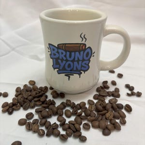 May include: A white ceramic coffee mug with the text "BRUNO LYONS" in blue graffiti-style lettering, a brown cigar graphic, and steam rising from the cigar. Coffee beans are scattered around the mug.