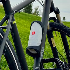 May include: A gray hybrid bicycle with black tires and a white stripe. A white and black speaker is mounted on the frame. A red taillight is attached to the seat post. The bike is on a grassy path.