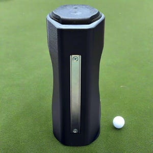 May include: A black golf club accessory with a silver accent and a white golf ball on a green surface. The accessory has a unique shape and a textured top.
