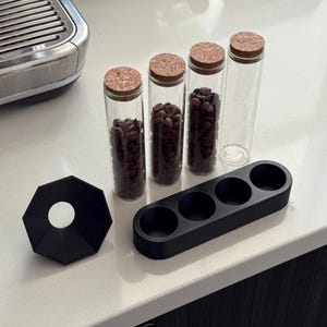 May include: A coffee station setup featuring four glass vials with cork stoppers, two filled with coffee beans. A black, octagonal base and a black tray with four circular indentations are also present. The items are on a white countertop.