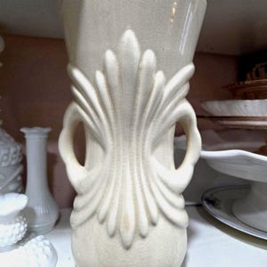 May include: Cream-colored ceramic vase with a flared top and a bulbous base. The vase features a stylized, symmetrical design with vertical lines and two loop handles. The surface has a crackle glaze, adding to its vintage aesthetic. The vase is displayed on a white shelf.