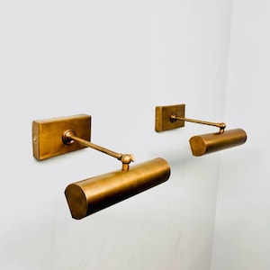 May include: Three brass-coloured wall-mounted picture lights. Each light features a rectangular base, an adjustable arm, and a cylindrical light fixture. They are mounted on a white wall, offering a warm, ambient glow.