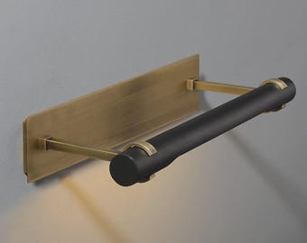 May include: A wall-mounted light fixture with a rectangular brass backplate and a cylindrical black light source. The fixture is supported by brass arms and is designed for indoor use. The light is turned on.